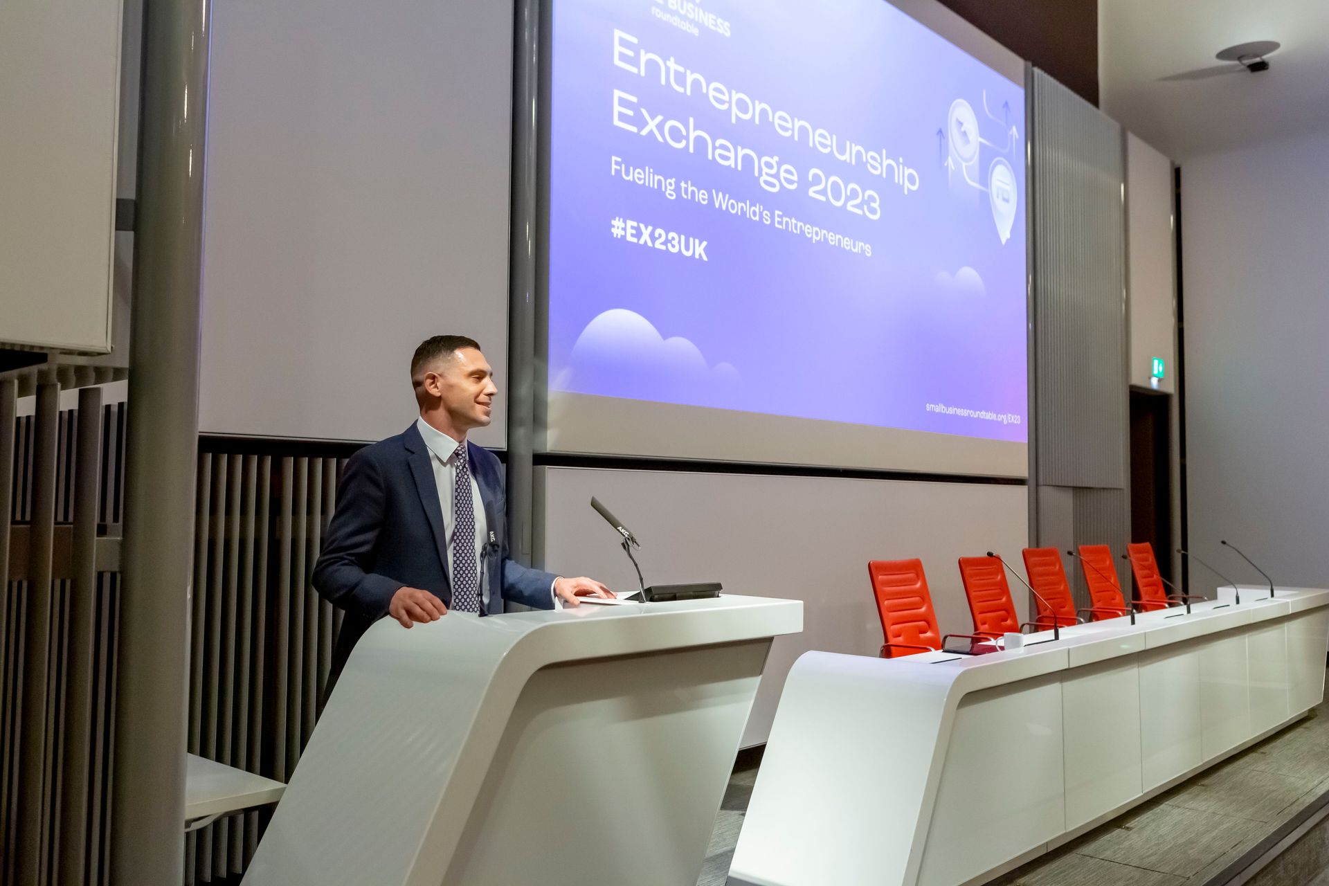 A man is standing at a podium giving a presentation in front of a large screen.