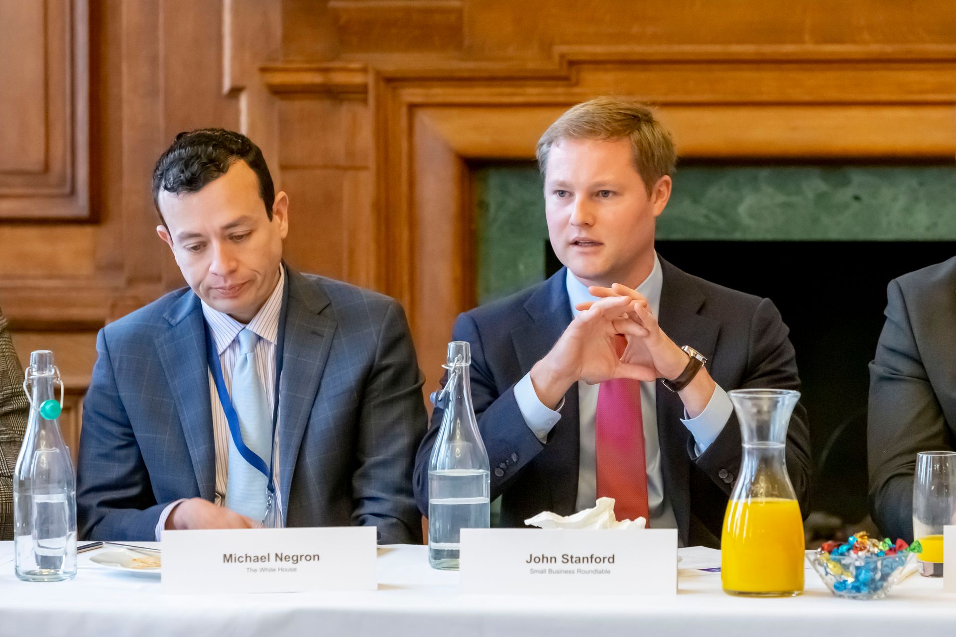 Two men in suits and ties are sitting at a table with bottles of water and orange juice.