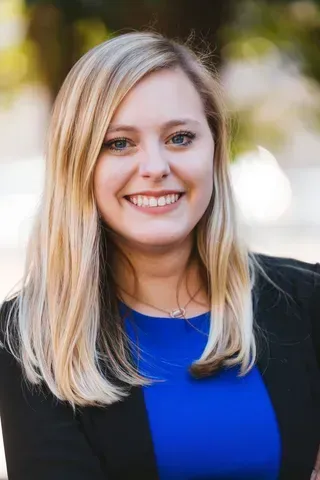 A woman in a blue shirt and black jacket is smiling for the camera.