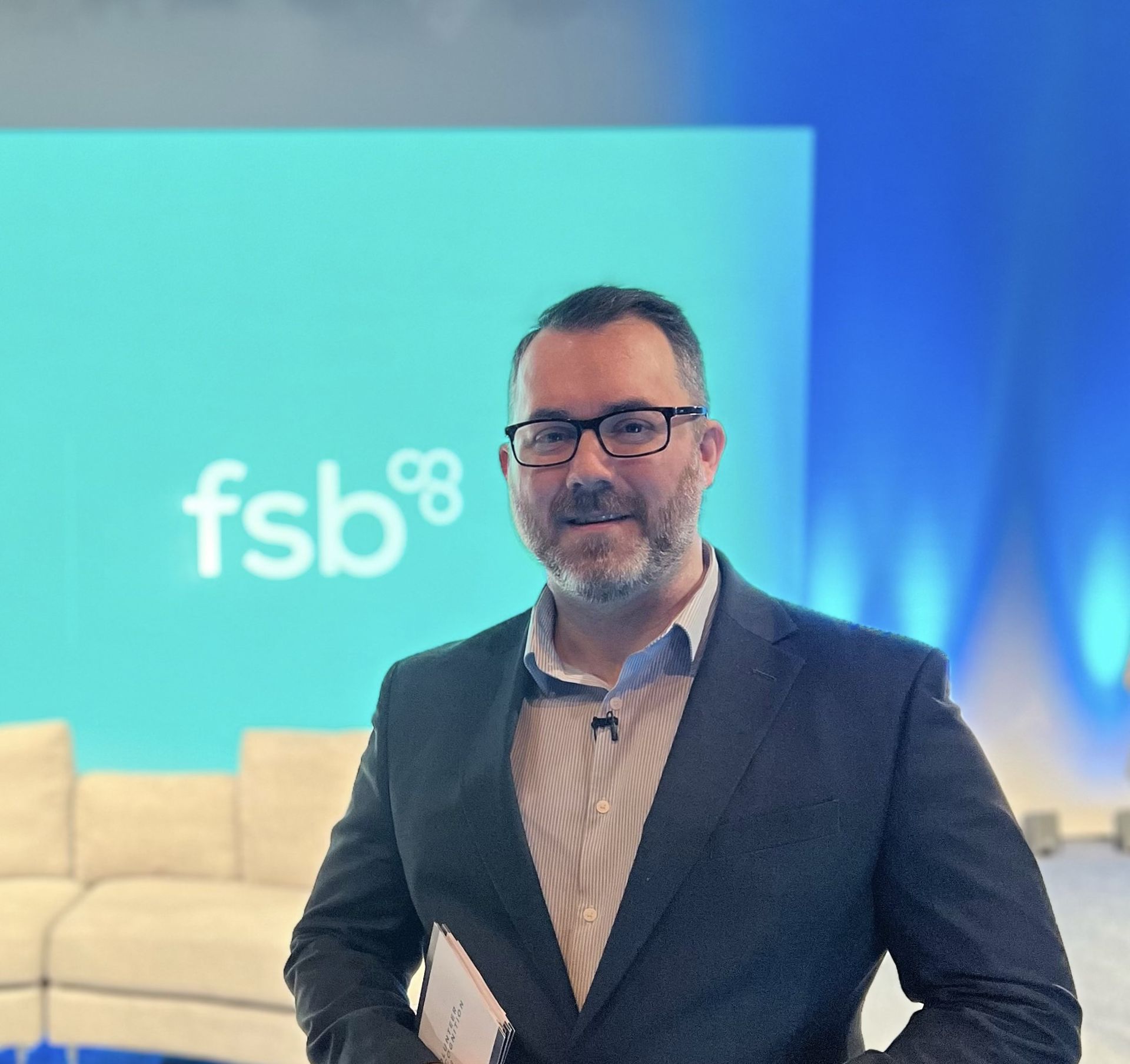 A man in a suit and glasses stands in front of a fsb logo