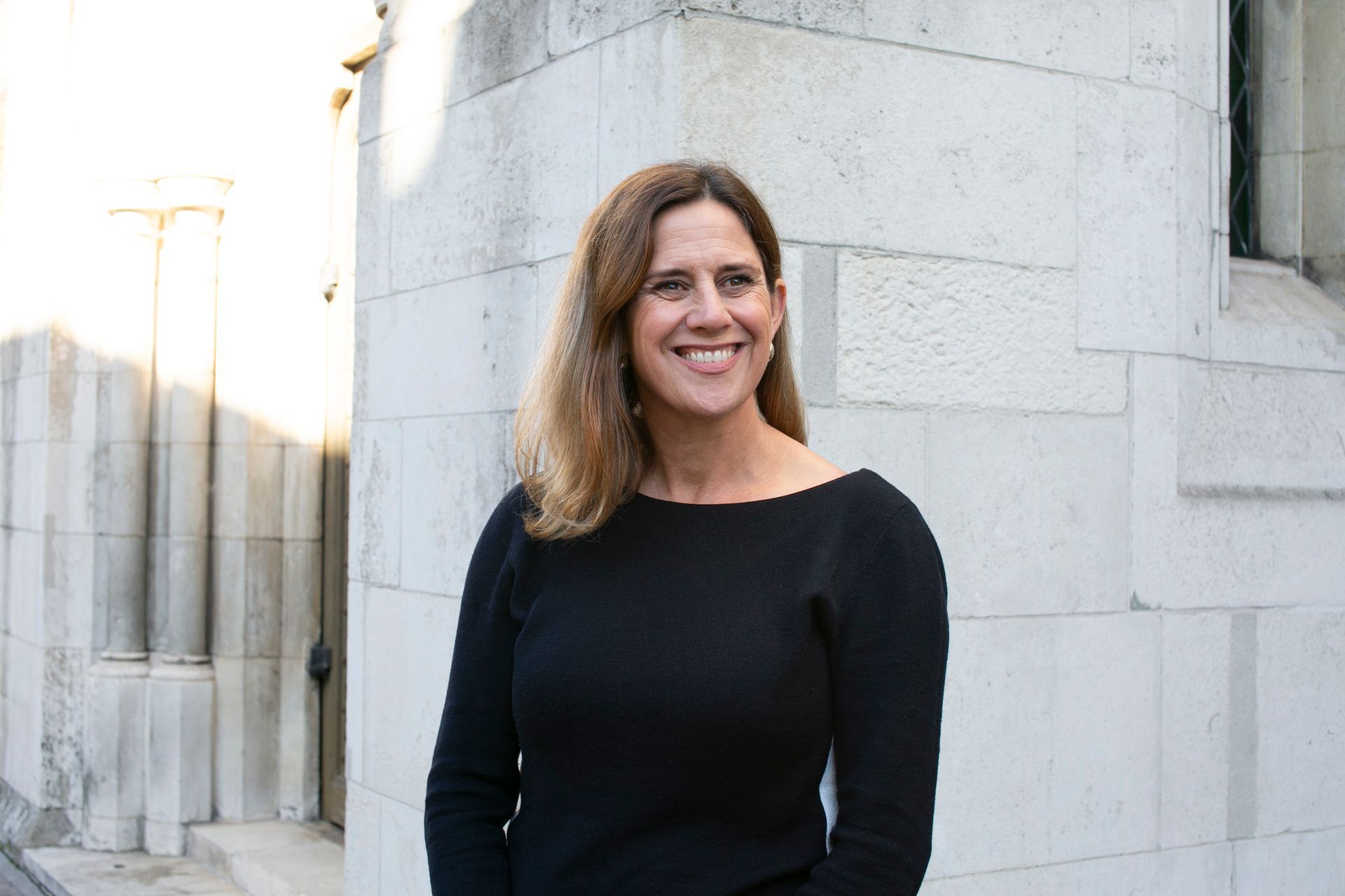 A woman in a black dress is standing in front of a brick wall and smiling.