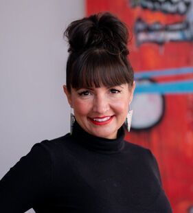 A woman wearing a black turtleneck and earrings is smiling in front of a painting.