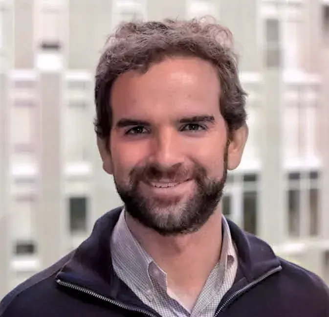 A man with a beard is smiling in front of a blurry background of buildings.