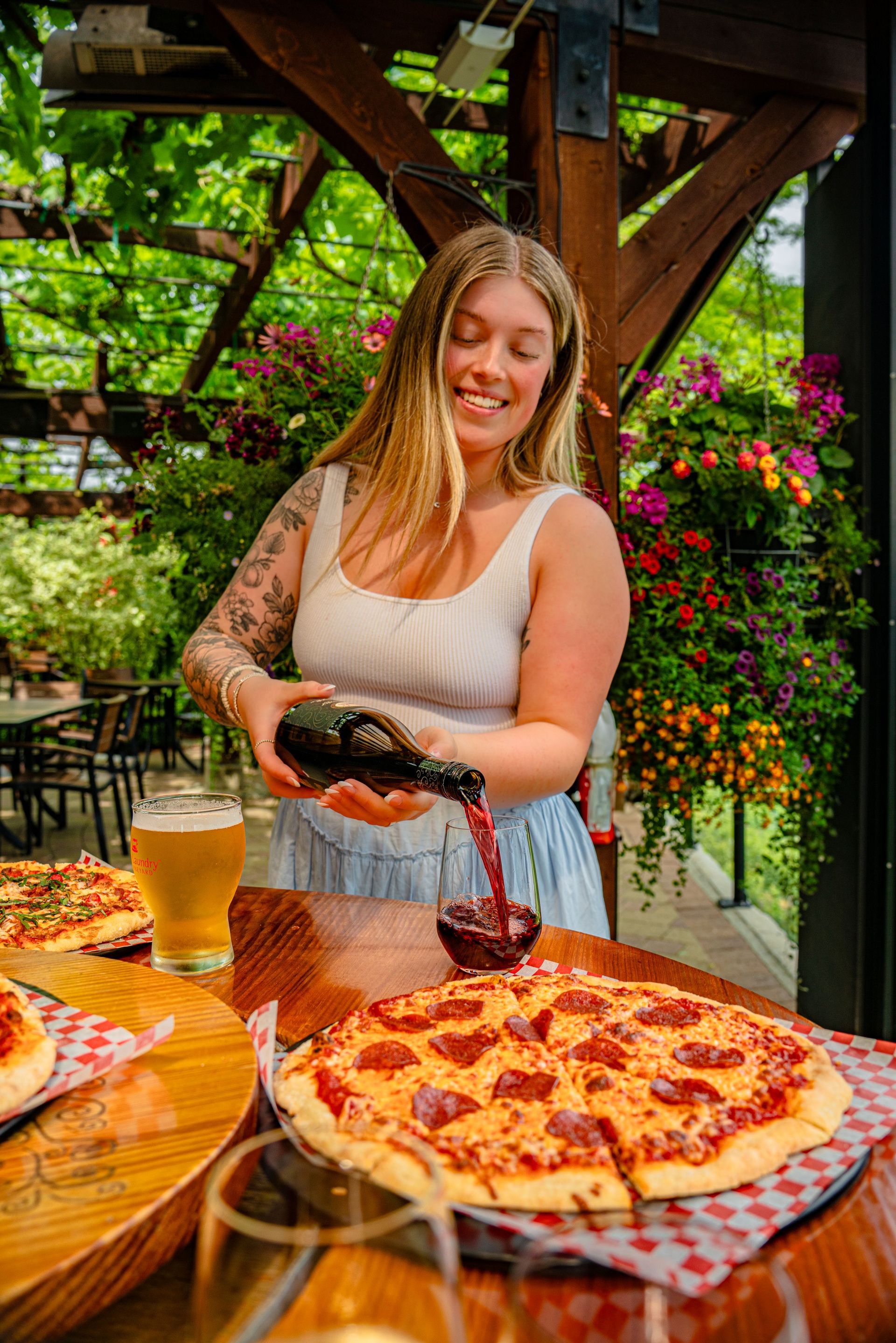 Woman pouring red wine, smiling, next to pizza and beer, outdoor patio.
