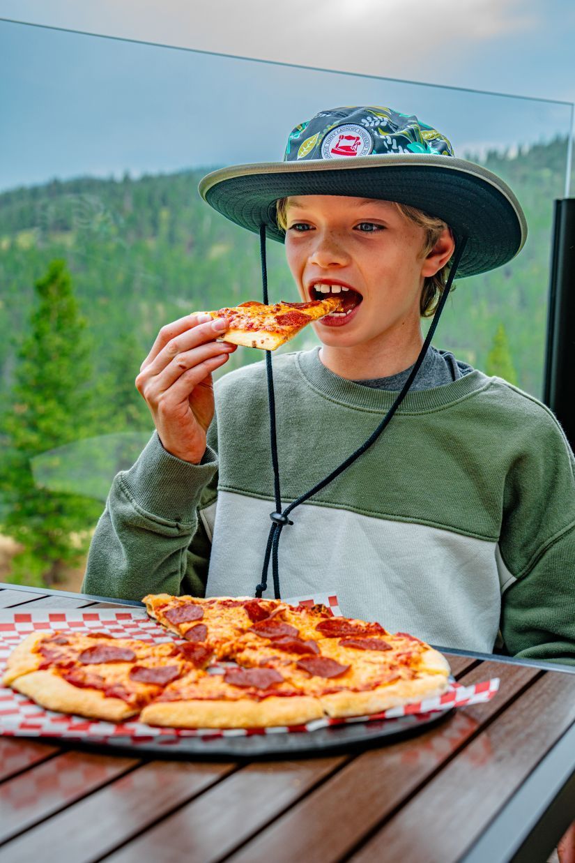 Boy eating a slice of pepperoni pizza outdoors, wearing a hat, enjoying the meal.