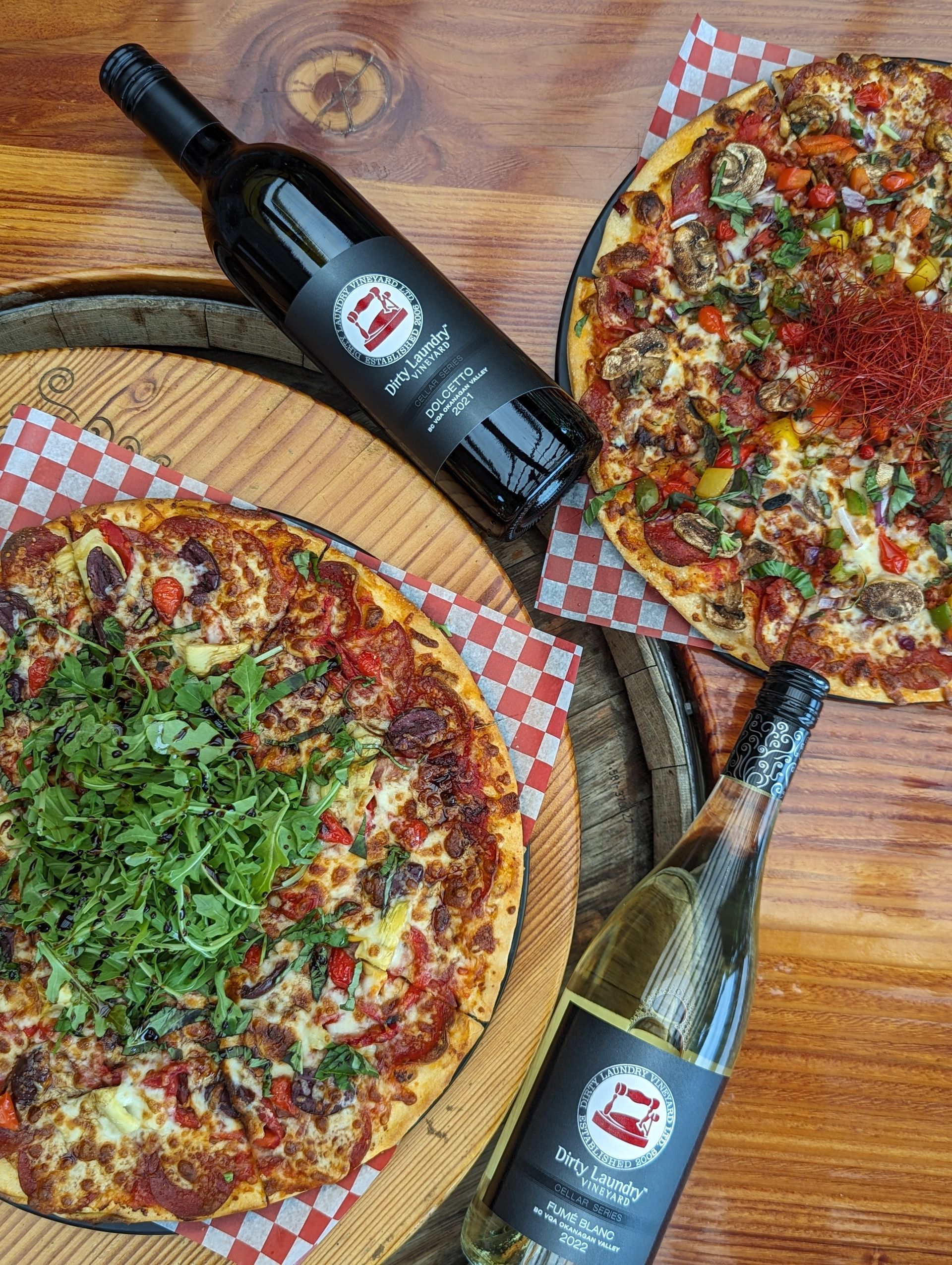 Three pizzas and two bottles of wine on a wooden table.