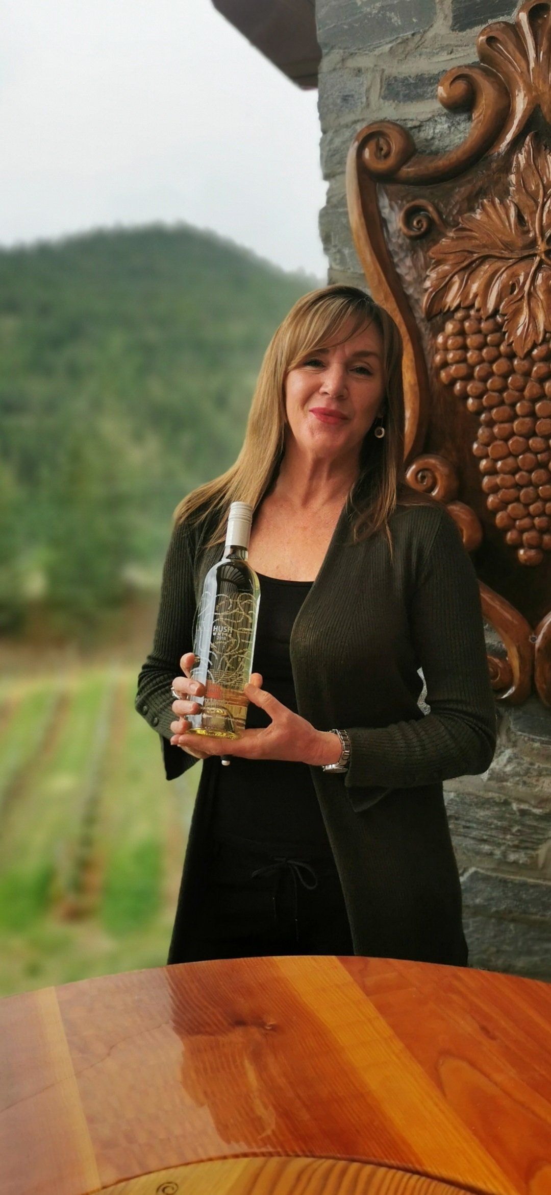 A woman is holding a bottle of wine in front of a wooden table.