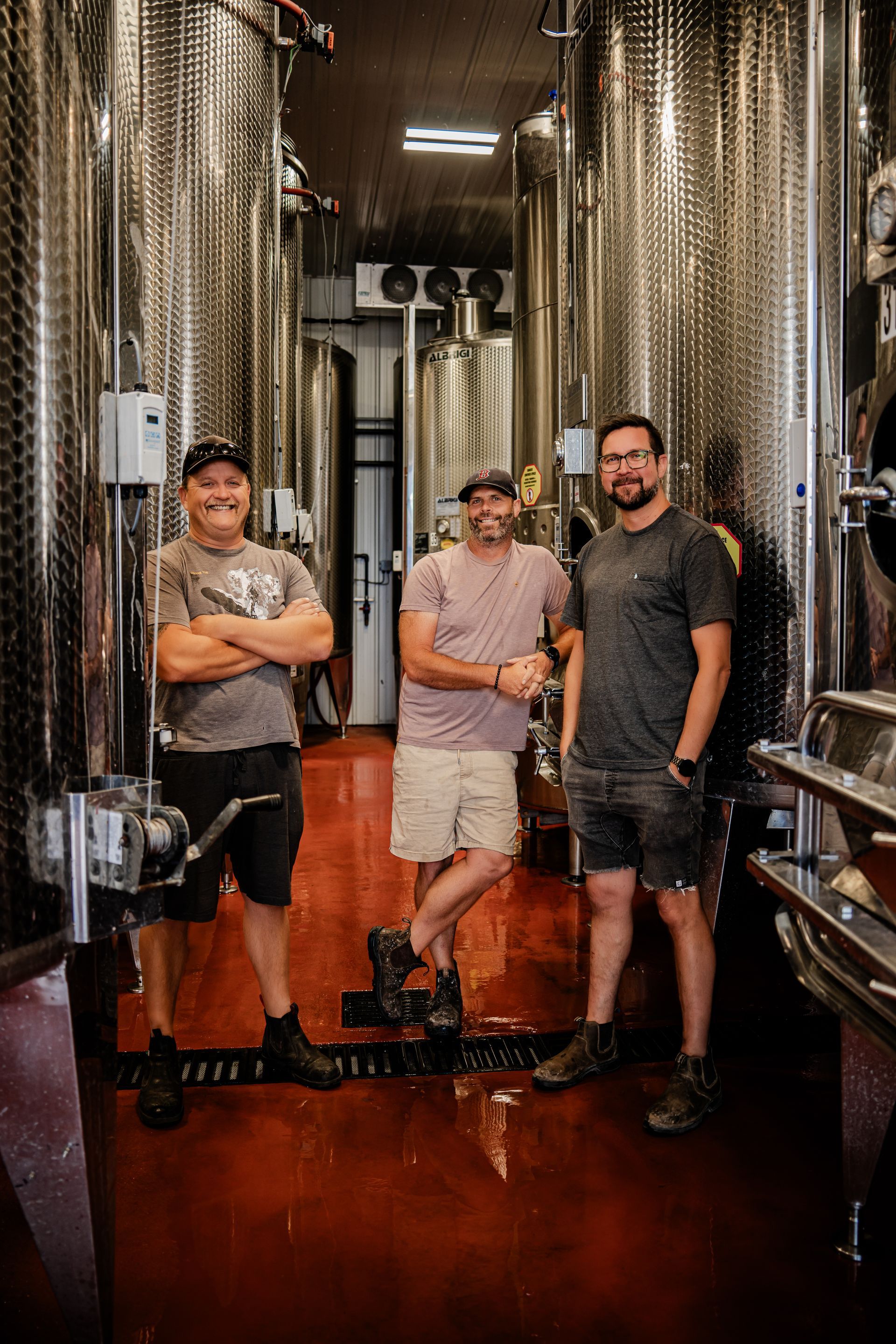 Three people in a winery, posing near large steel tanks. Two men are smiling, one with arms crossed.
