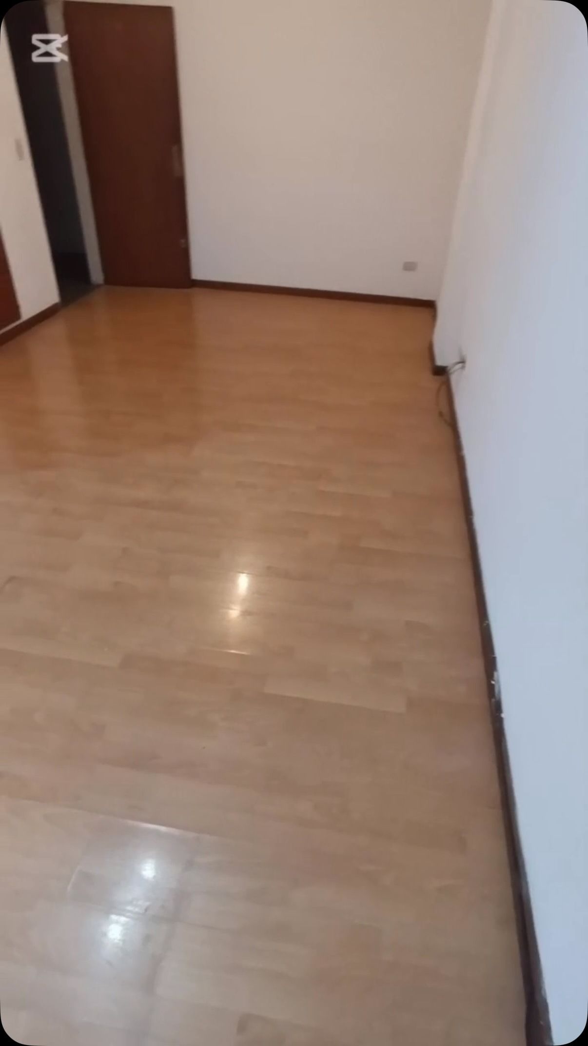 Habitación vacía con suelo de madera clara, paredes blancas y una puerta marrón en la esquina.