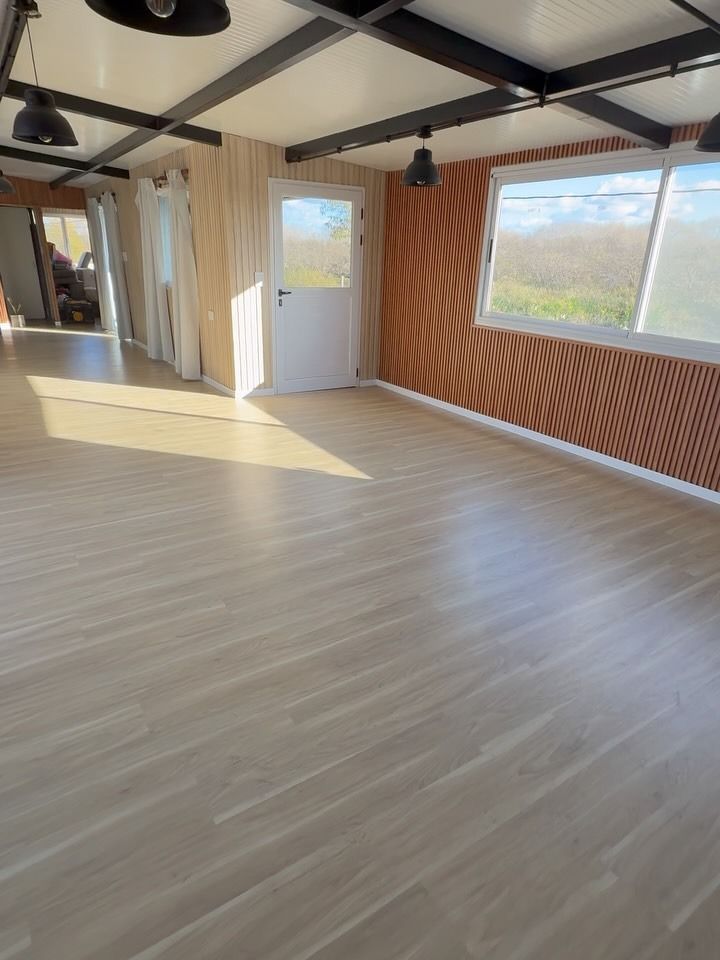 Habitación vacía con suelo de madera clara, vigas a la vista, grandes ventanales y una puerta blanca.
