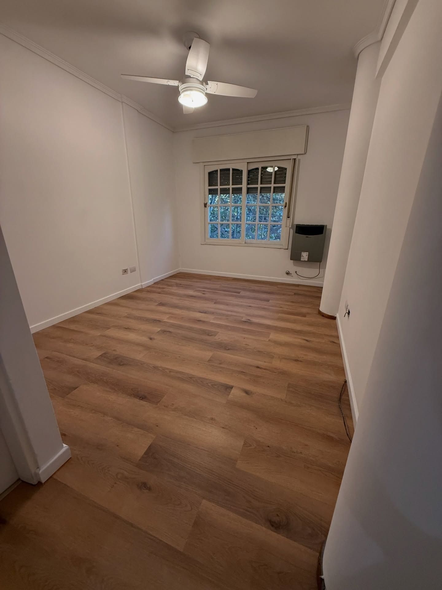Habitación vacía con suelo de madera, paredes blancas, ventilador de techo y ventana con persianas.