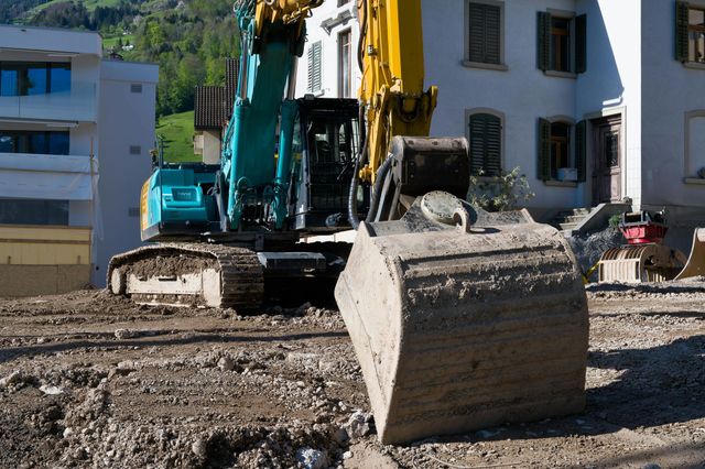 A construction site with an excavator in front of a building. The excavator is teal and yellow.
