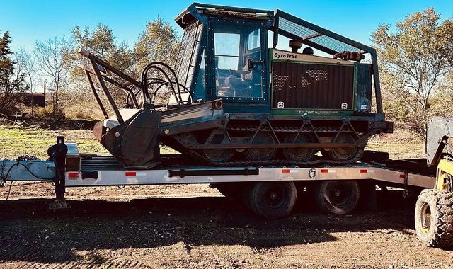 A large, dark-green forestry machine on a trailer, parked outdoors on a sunny day.