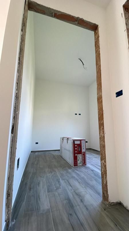 Stanza vuota e non finita, con pareti bianche, pavimento grigio effetto legno e un telaio di porta in primo piano