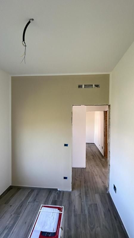 Stanza bianca vuota con pavimento in legno grigio e una porta che dà su un corridoio; si vedono i cavi a vista sul soffitto.