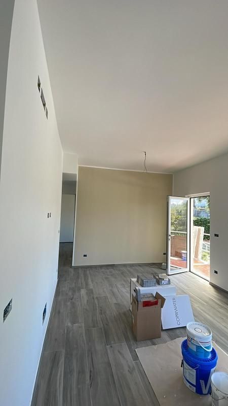 Stanza stretta in ristrutturazione, con pavimento grigio, parete beige, porta a vetri aperta e vernici a terra