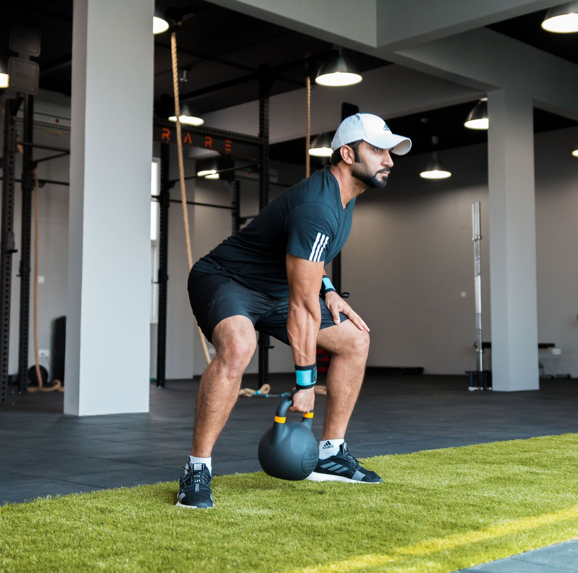 Man lifting a kettlebell in a gym in Lynden, WA, standing on green turf and wearing black athletic gear and a white cap.