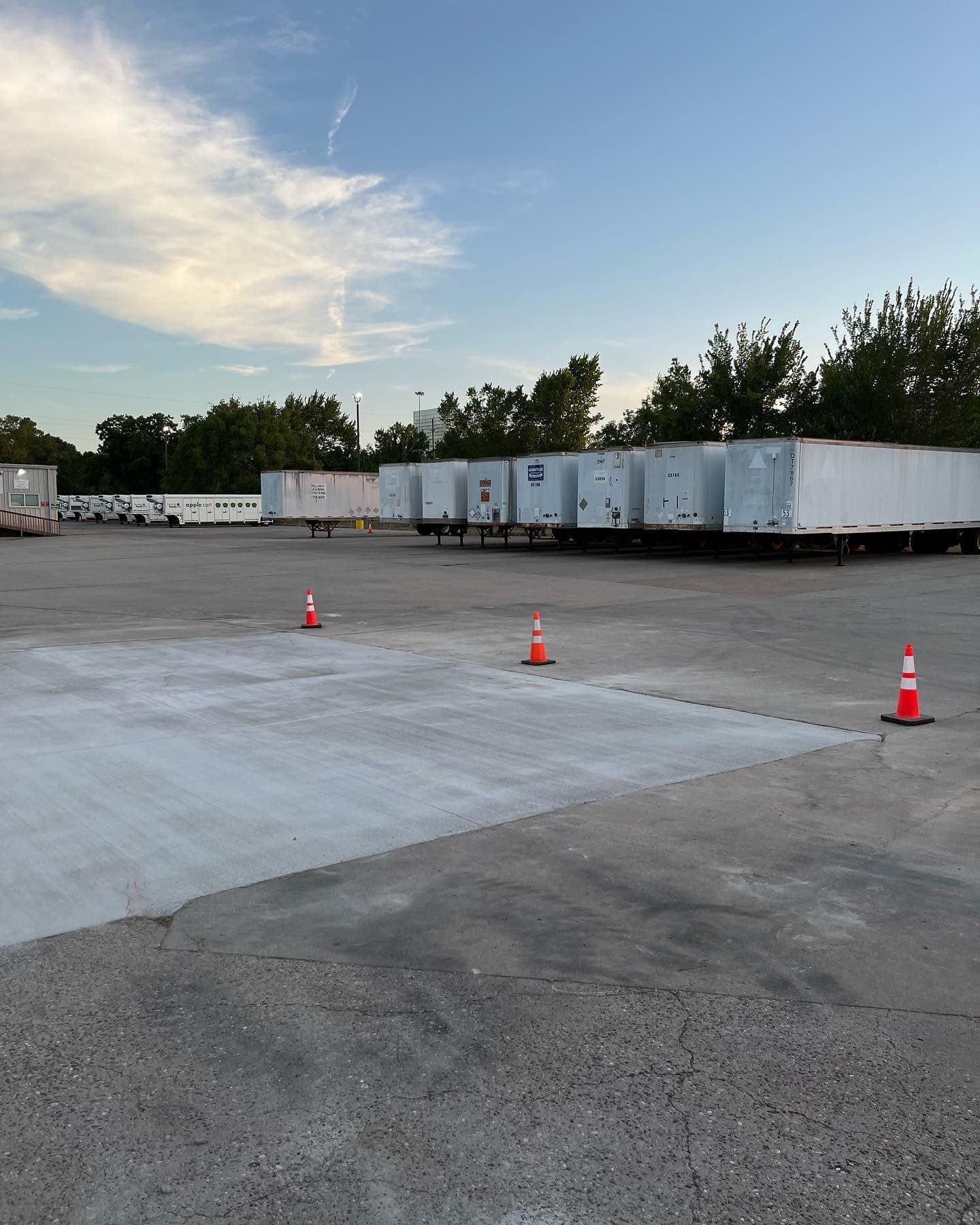 Empty parking lot with several parked semi-truck trailers under a blue sky. Orange cones mark areas on the ground.