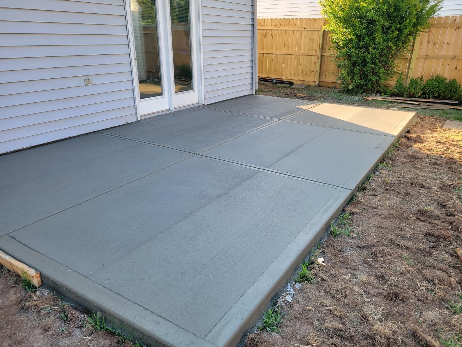 Newly poured concrete patio next to a house with a wooden fence in the background.