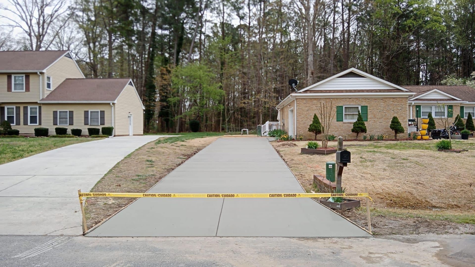 A newly poured concrete driveway is blocked by yellow caution tape, flanked by two houses.