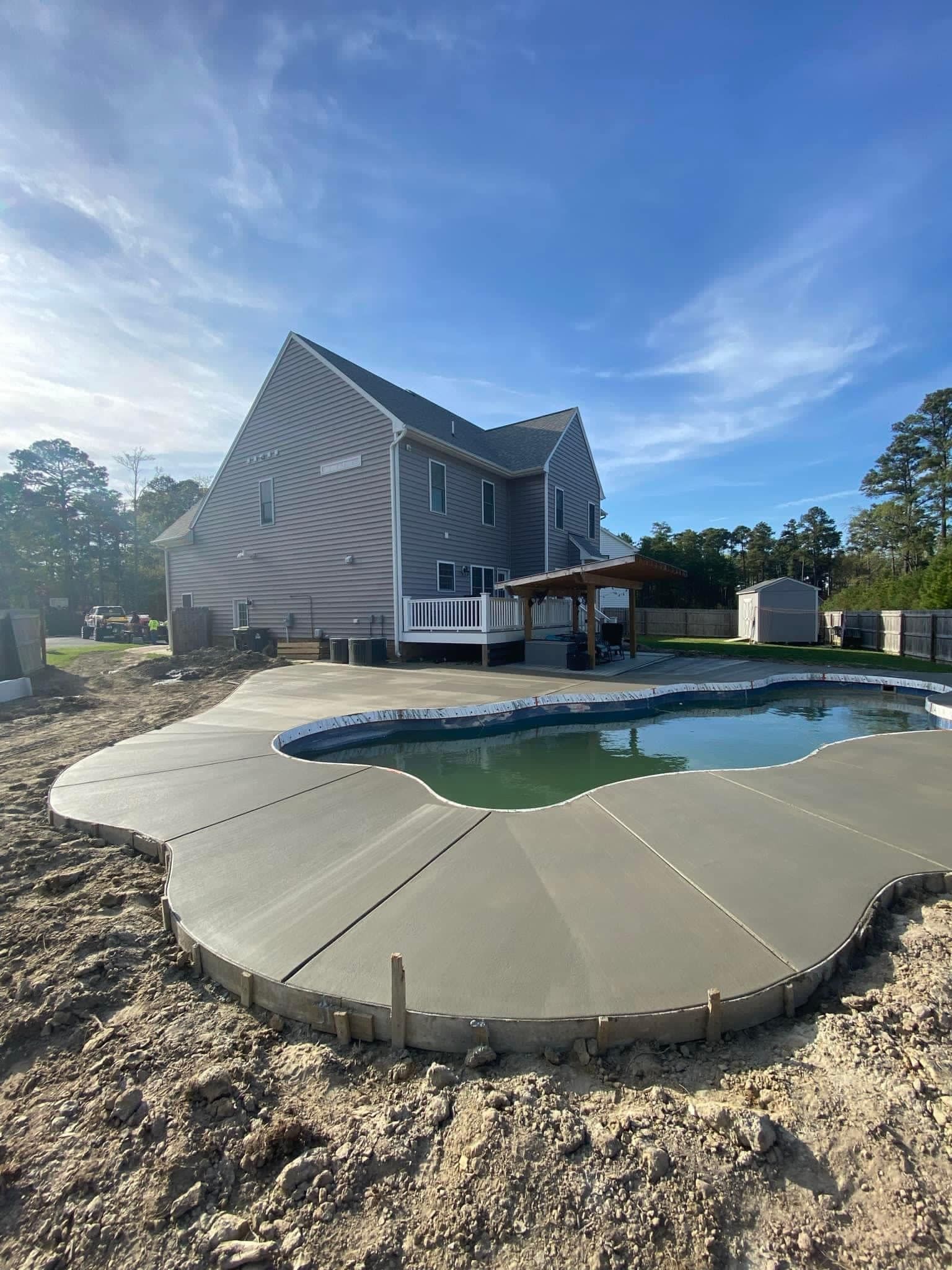 Pool construction with concrete deck, house backdrop, blue sky.