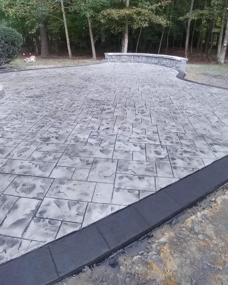 Stamped concrete patio with gray stone pattern, black border, and stone wall in a wooded setting.