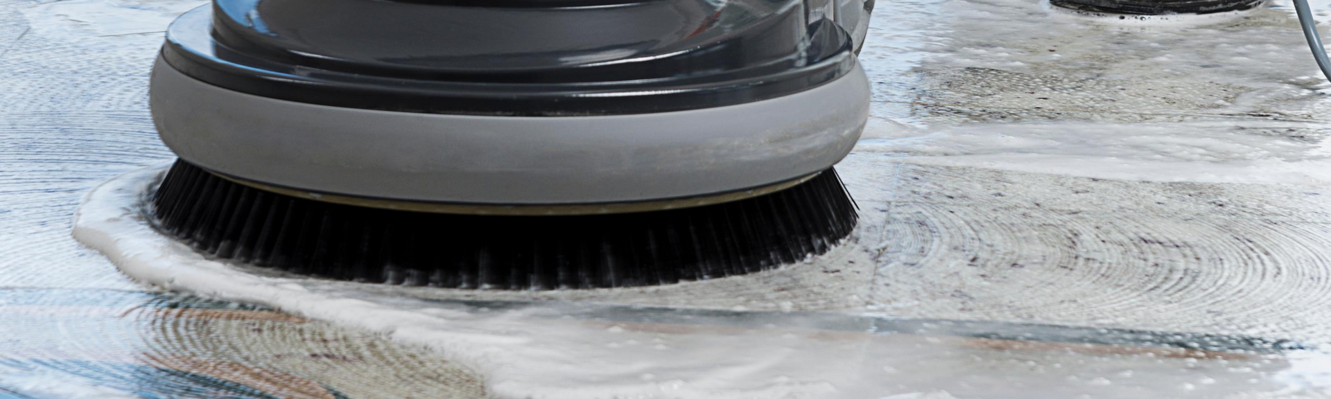 Floor scrubber cleaning a wet concrete surface, with black bristles and gray housing