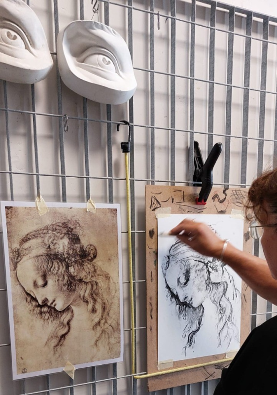 Persona che disegna un ritratto, confrontandolo con un disegno di riferimento. Due calchi in gesso di occhi sono appesi sopra.