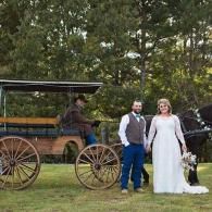 Bride and groom posing beside a horse-drawn carriage in a grassy outdoor setting