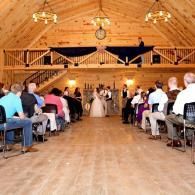 Wedding ceremony in a wooden barn, guests seated on both sides of the aisle as the couple stands at the front
