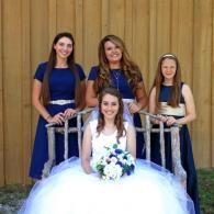 Four women in blue dresses pose at a wooden wall, with one seated in a white ball gown holding a bouquet.