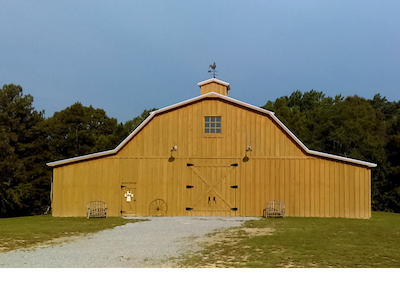 Yellow wooden barn with a gravel driveway and trees under a cloudy sky