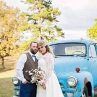 Newlyweds pose beside a blue vintage truck outdoors in a sunny autumn setting