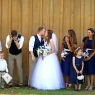 Wedding couple kissing with bridal party standing beside them outdoors against a wooden wall