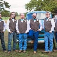 Five men in matching vests and bow ties stand in front of a blue truck outdoors.