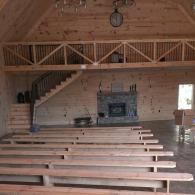 Rustic chapel interior with wooden pews, staircase, loft balcony, and stone fireplace/altar at the front