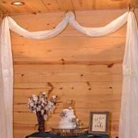 White draped fabric on a wooden wall above a table with flowers, cake, and framed decor
