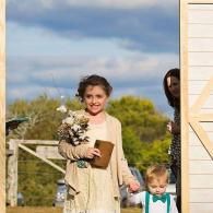 Two children at a doorway, one holding flowers and a small pot, with a smiling adult partially visible behind them.