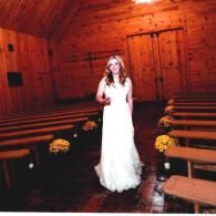 Bride in a white gown standing in a wooden chapel aisle with yellow flowers on the pews.