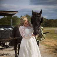 Bride in a white lace dress embracing a black horse beside a carriage under a cloudy sky