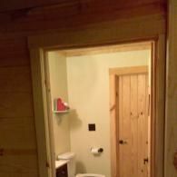 Small bathroom doorway with a toilet, shower, and wooden door visible inside.