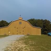 Yellow barn with a curved roof on a gravel drive, set against trees under a blue sky