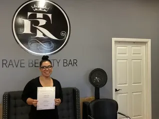 Woman with glasses under logo holding certificate | Sylmar, CA | Rave Beauty Bar