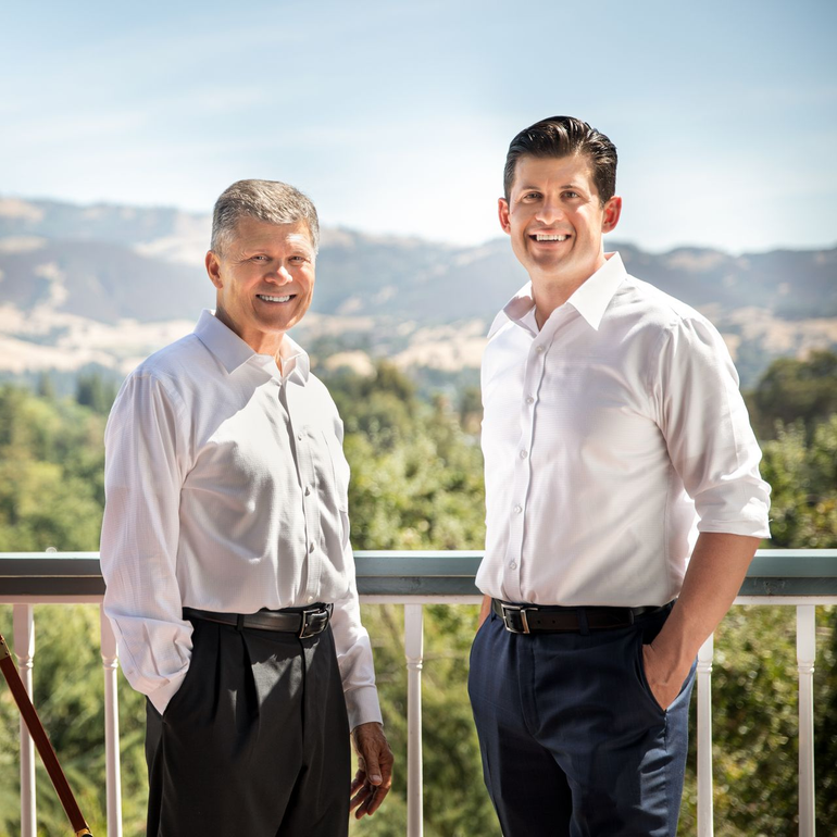 Two men in white shirts and belts stand outside smiling, with a nature backdrop.