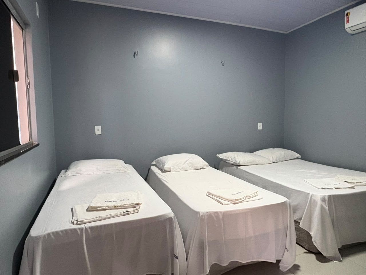 Quarto pequeno com três camas de solteiro cobertas com lençóis brancos e toalhas dobradas, paredes cinzentas e uma janela.