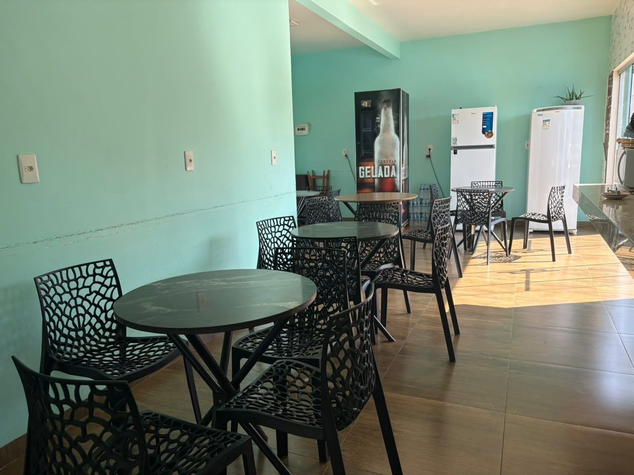 Sala de jantar vazia com cadeiras pretas e mesas redondas contra paredes verde-menta e piso de madeira.