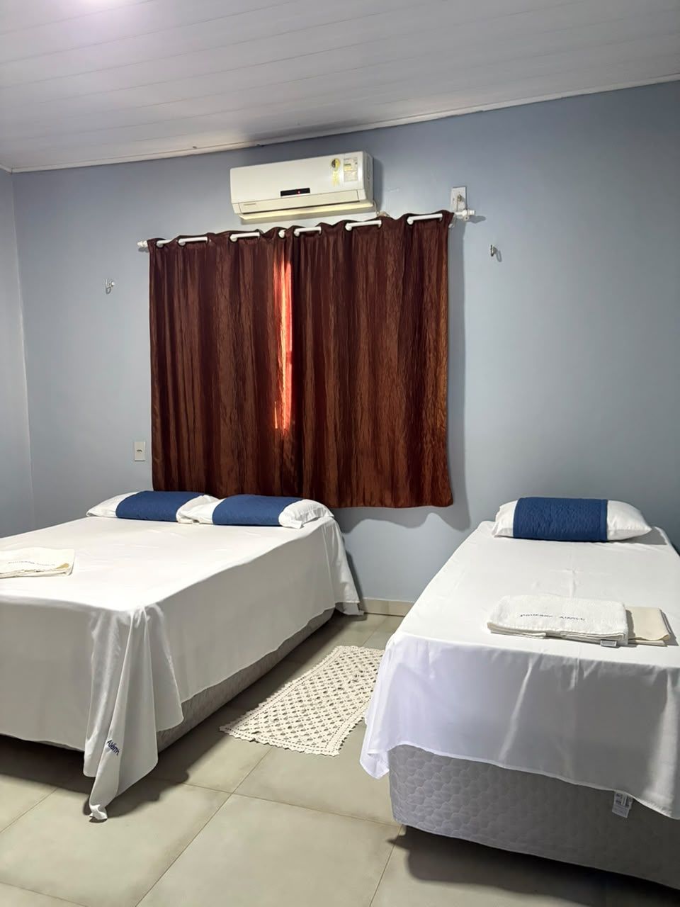 Duas camas de solteiro em um quarto simples com cortinas, ar-condicionado e toalhas dobradas.