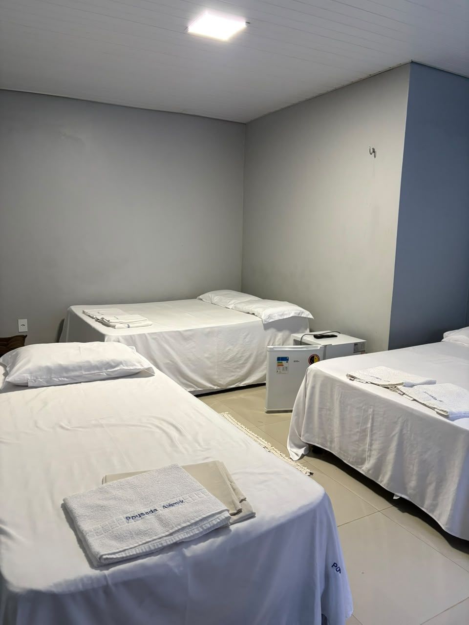 Quarto pequeno com quatro camas de solteiro desarrumadas e lençóis brancos sob uma parede cinza lisa.
