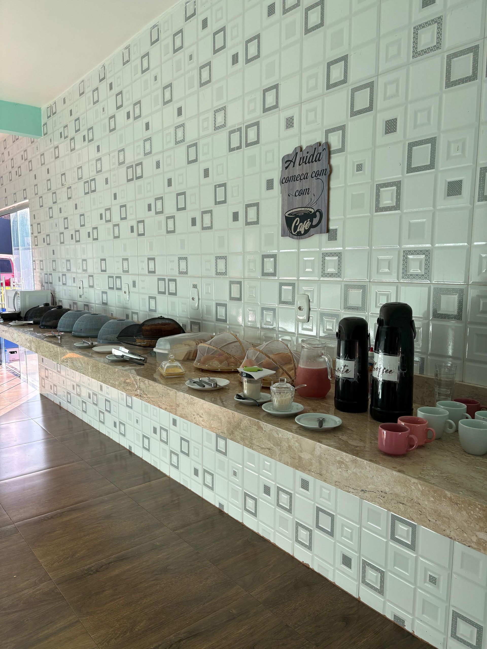 Balcão de buffet com bandejas de comida, pratos e dispensadores de bebidas contra uma parede revestida com azulejos estampados.