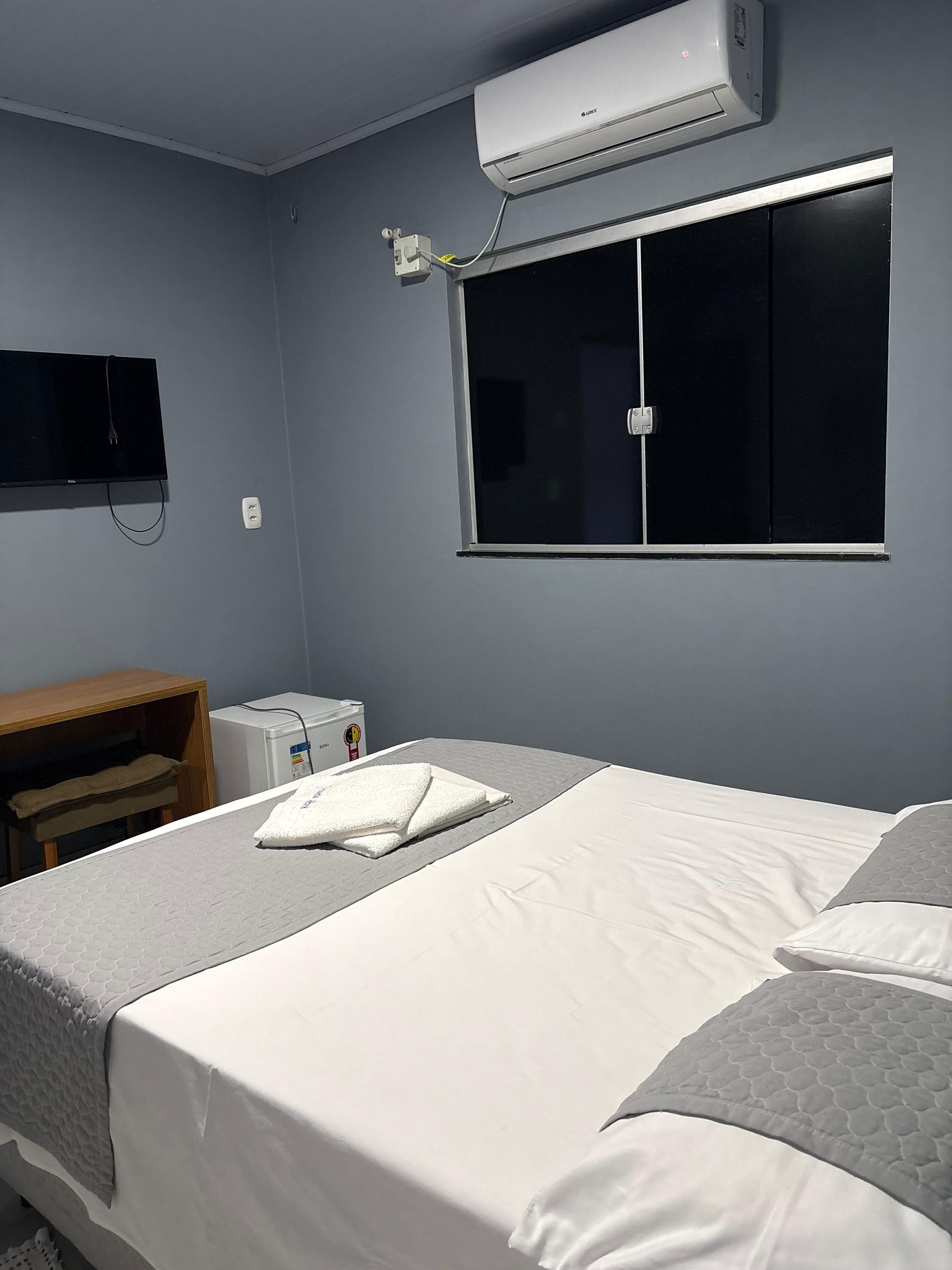Quarto pequeno com cama, TV de parede, ar condicionado e janela com persianas escuras.